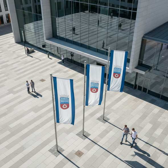 Draufsicht auf den Rathausplatz: drei Hissfahnen in Blau-Weiß, Personen mit langen Schatten.