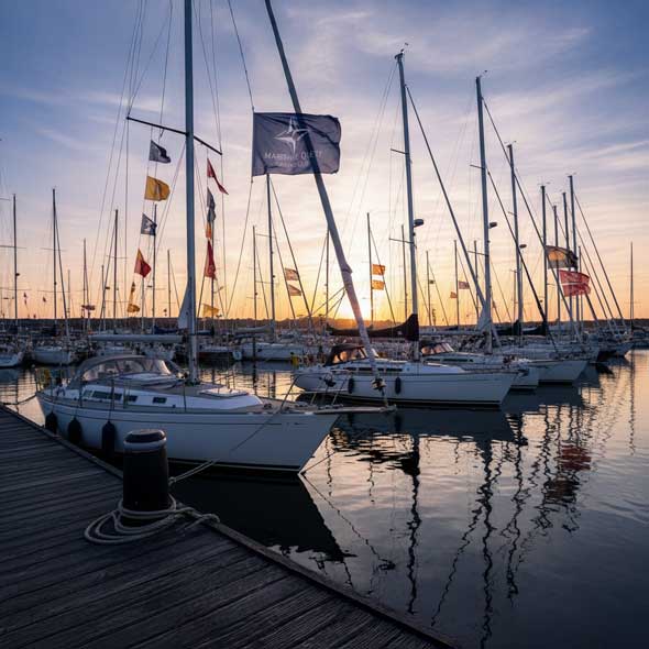 Mehrere Segelboote im Yachthafen bei Sonnenuntergang, an den Masten wehen Velumflag Longlife Flaggen mit Vereinsdesigns; ruhige Abendstimmung, klare Spiegelungen im Wasser.