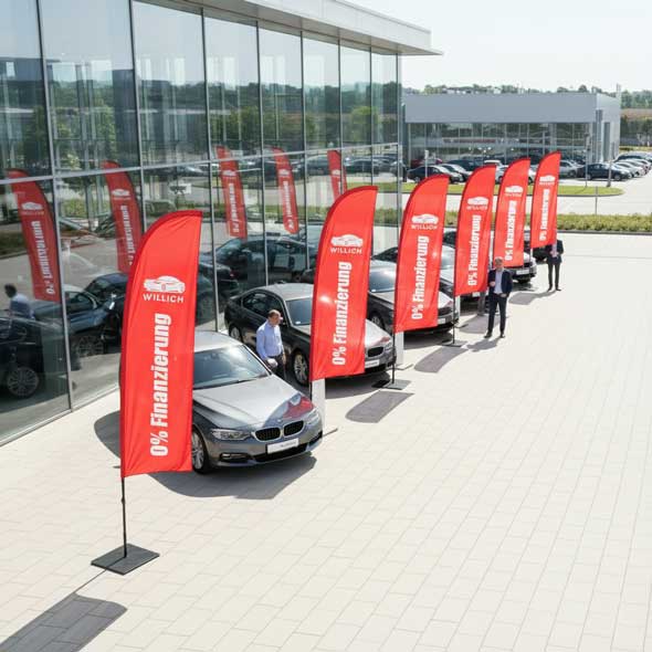 Rote Beachflags mit Aufdruck „0 % Finanzierung“ vor einem Autohaus mit Fahrzeugen – auffällige Werbefahnen für Autohändler und Verkaufsaktionen im Außenbereich.